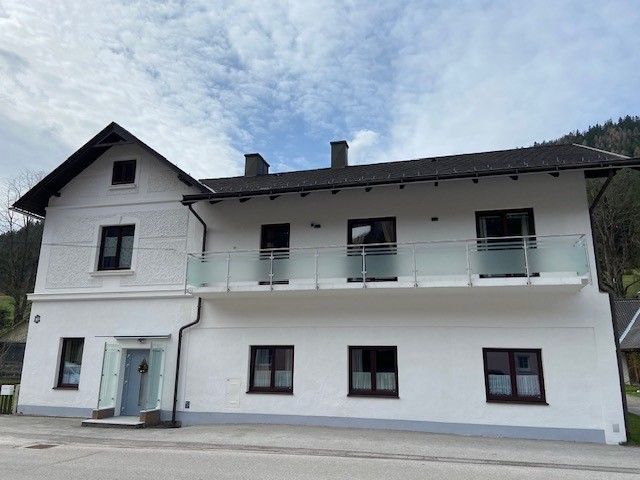 Zweistöckiges weißes Haus mit Balkon und blauen Himmel im Hintergrund.