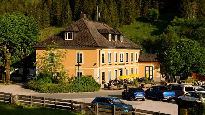 Ein gelbes Wirtshaus mit Terrasse und Sonnenschirmen, umgeben von Bäumen und Autos, vor einem bewaldeten Hügel.