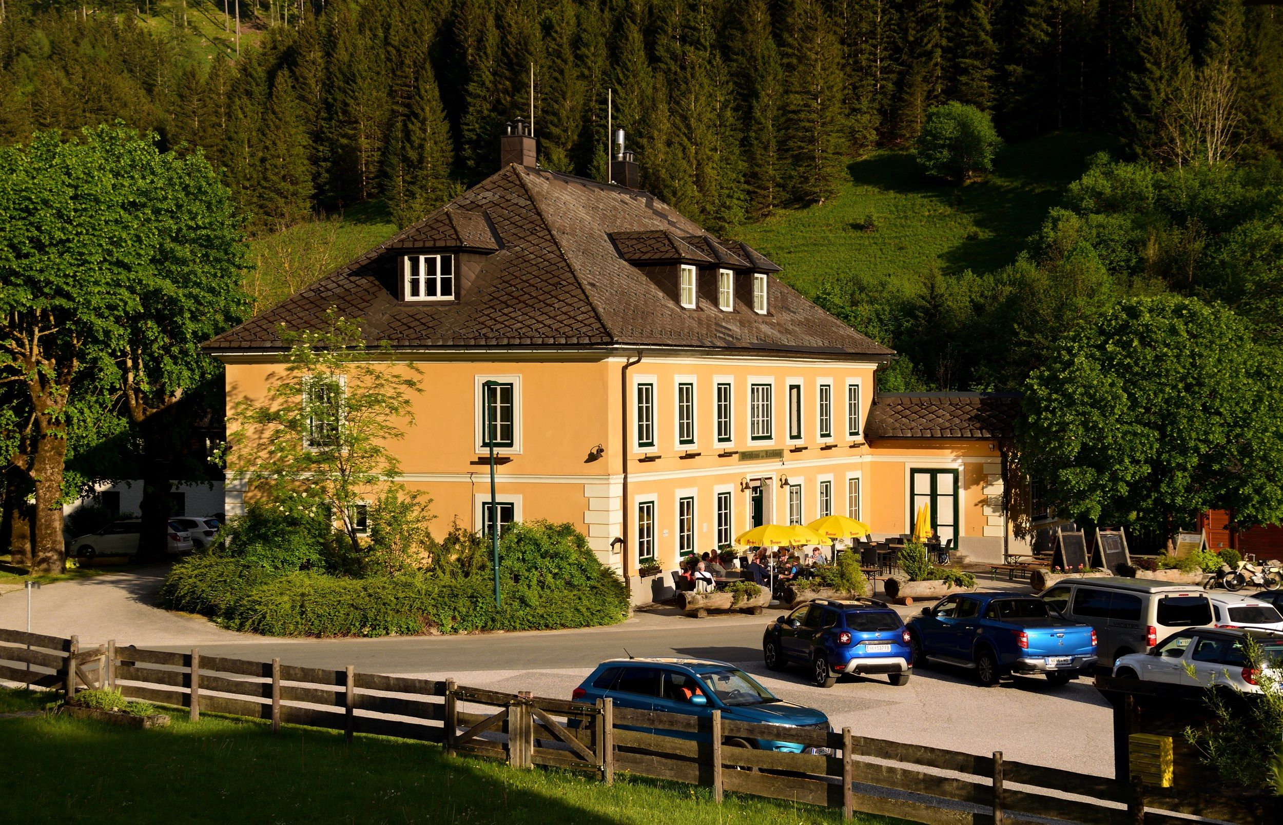 Ein gelbes Wirtshaus mit Terrasse und Sonnenschirmen, umgeben von Bäumen und Autos, vor einem bewaldeten Hügel.