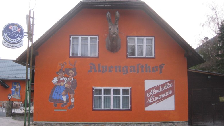 Orange gestrichenes Geb&auml;ude mit der Aufschrift 'Alpengasthof', Wandmalerei von tanzendem Paar und Almdudler-Werbung.