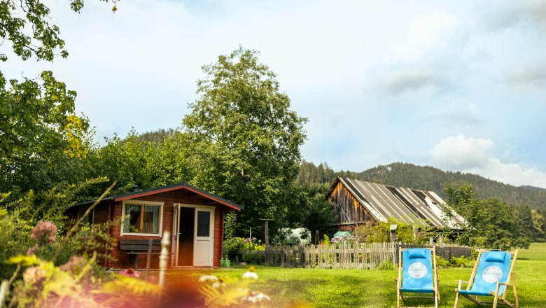 Ein idyllischer Garten mit einem kleinen Holzhaus, zwei blauen Liegest&uuml;hlen und einem alten Scheunengeb&auml;ude im Hintergrund.