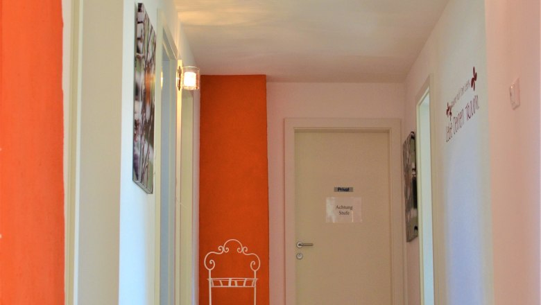 Ein Flur mit Holzboden, wei&szlig;en W&auml;nden und einer orangefarbenen Wand. An der Wand h&auml;ngt ein Schild mit der Aufschrift 'Achtung Stufe'.