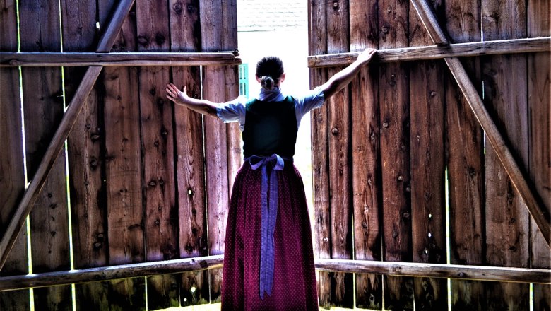 Eine Frau in traditioneller Kleidung &ouml;ffnet gro&szlig;e Holzt&uuml;ren nach au&szlig;en.