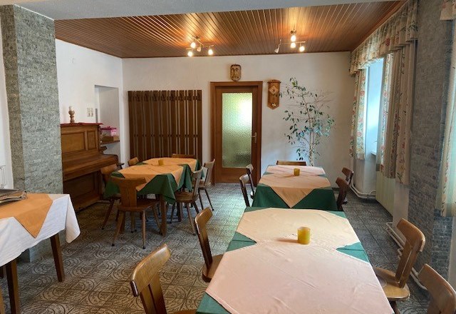 Ein gem&uuml;tlicher Fr&uuml;hst&uuml;cksraum mit Holztischen, St&uuml;hlen und einem Klavier. Die Tische sind mit Tischdecken gedeckt, und es gibt Fenster mit Vorh&auml;ngen.